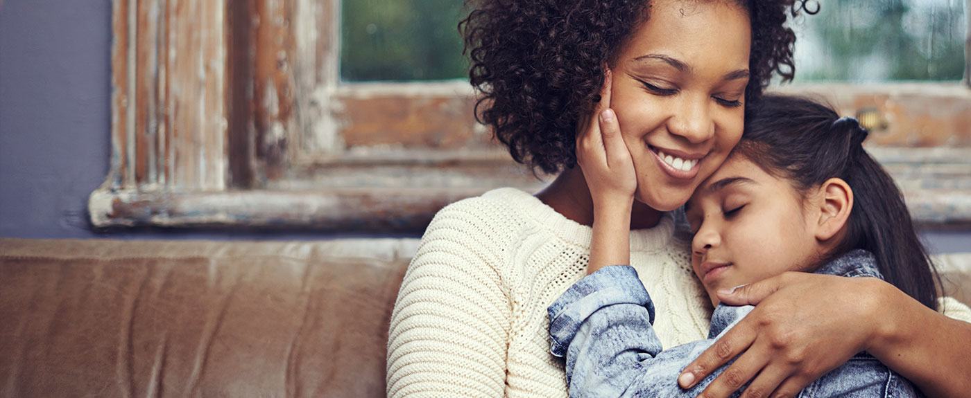 Mother and young daughter sitting on a couch hugging.